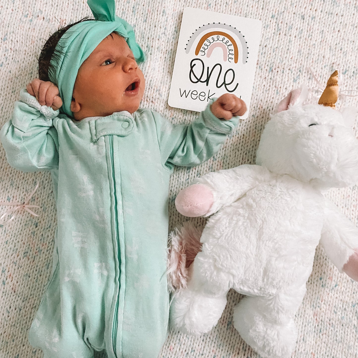 baby lying on a blanket with the one week old card next to her