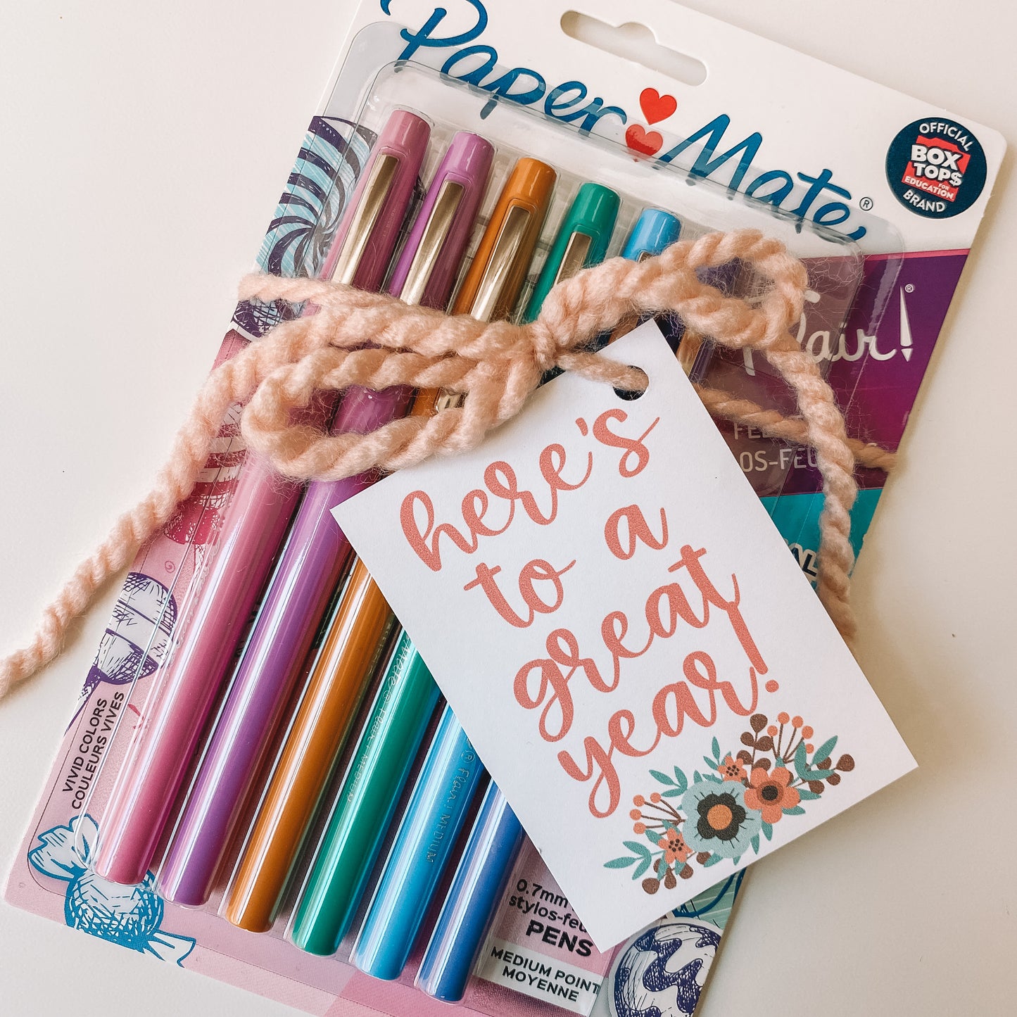 Here's to a great year gift tag has florals decorating the bottom of the tag. The tag is wrapped around a pack of papermade pens with a ribbon.