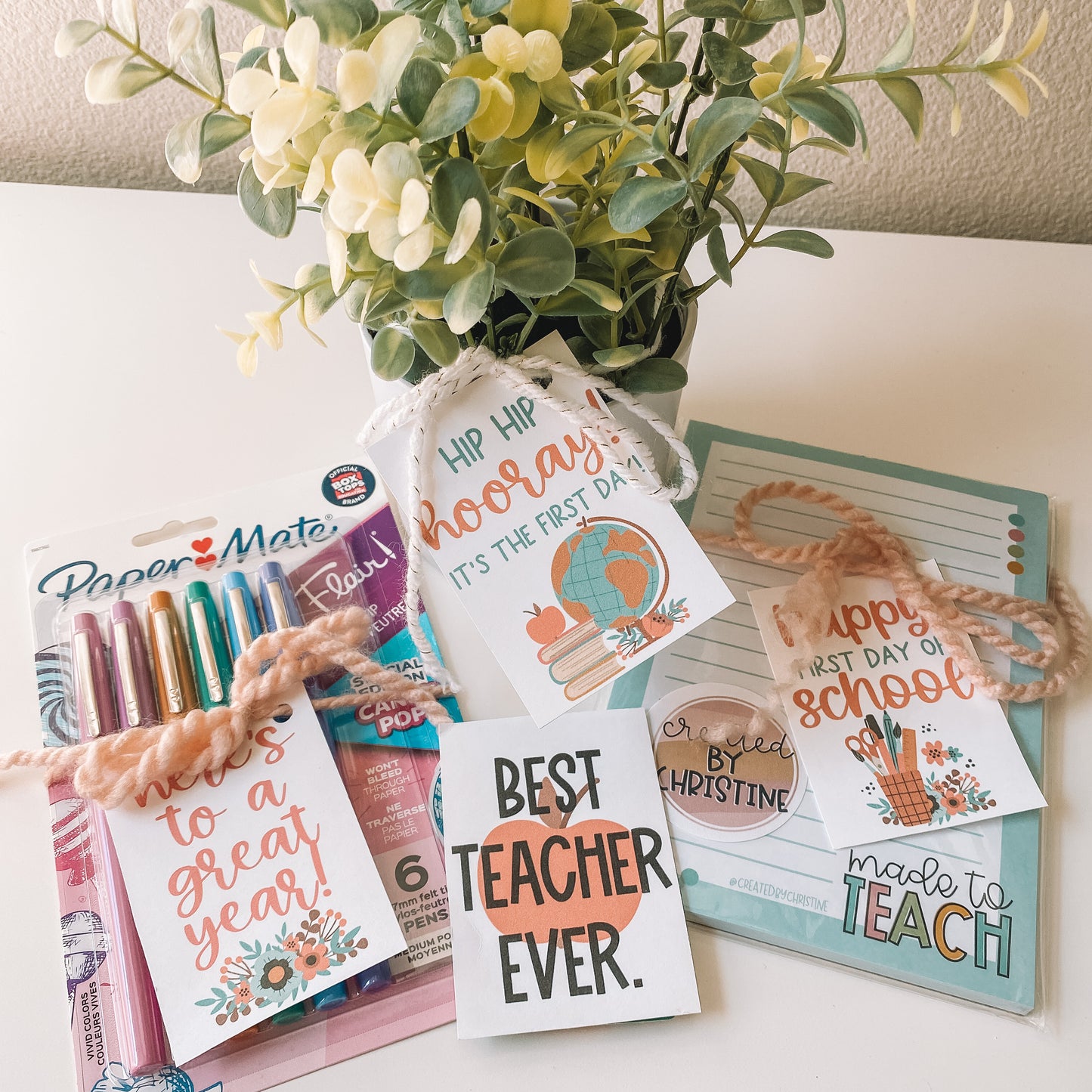 Gift tags attached with ribbon to a pack of papermade flair pens, a succulent plant, a teacher notepad, and a gift card.