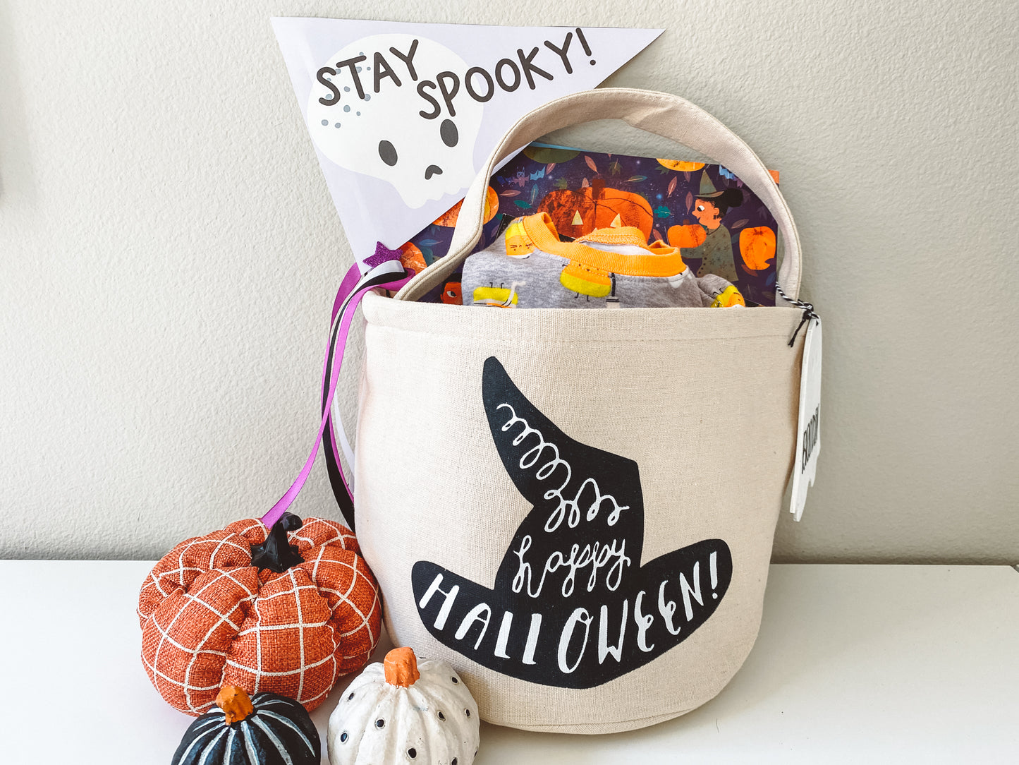 Boo Basket with books and pjs inside. Stay Spooky flag placed inside. Flag is light purple with black text and a cute white skull in the background. Three pumpkins are in front of the basket.