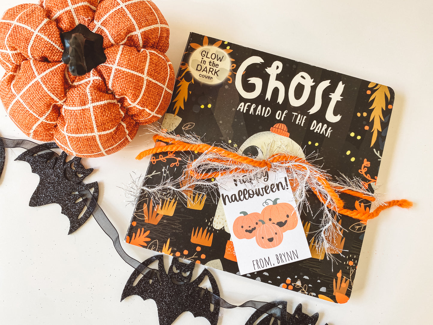 Halloween board book with ribbon wrapped around it and a happy halloween gift tag featuring three orange pumpkins and the gift giver's name at the bottom. Orange pumpkin and bat garland complete the flat lay.