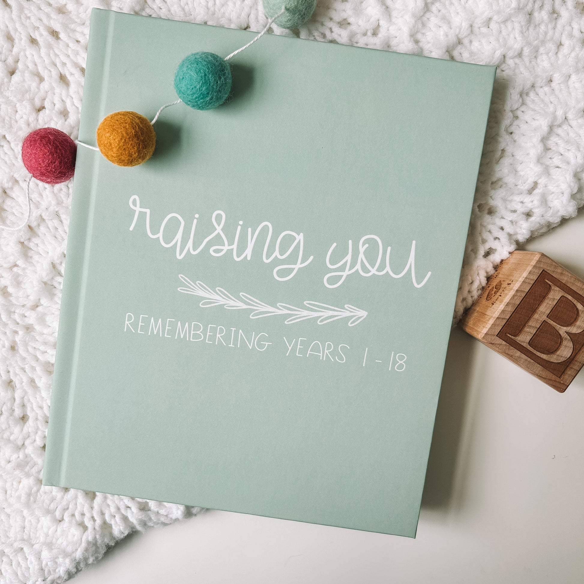 Green childhood memory book titled Raising You Remembering Years 1-18 in white text