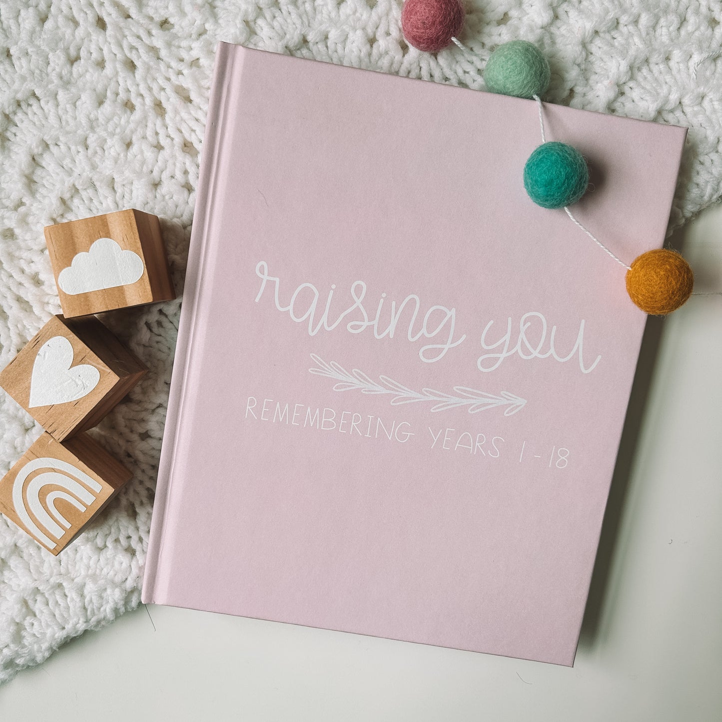 Pink childhood memory book titled Raising You Remembering Years 1-18 in white text