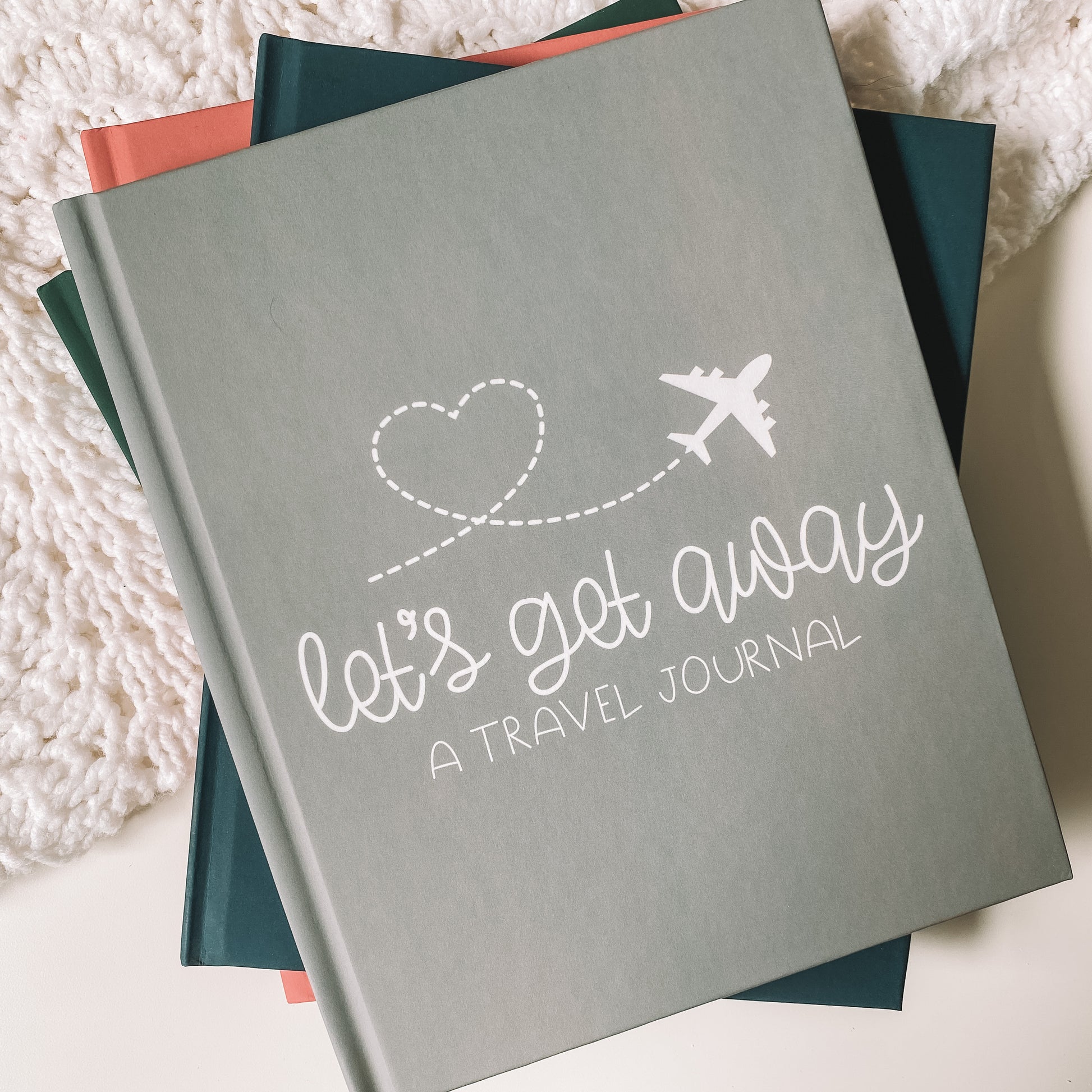 Keepsake travel journal with a soft, matte grey hardcover. White text reads Lets Get Away A Travel Journal with a graphic of a white airplane leaving a heart trail behind it.