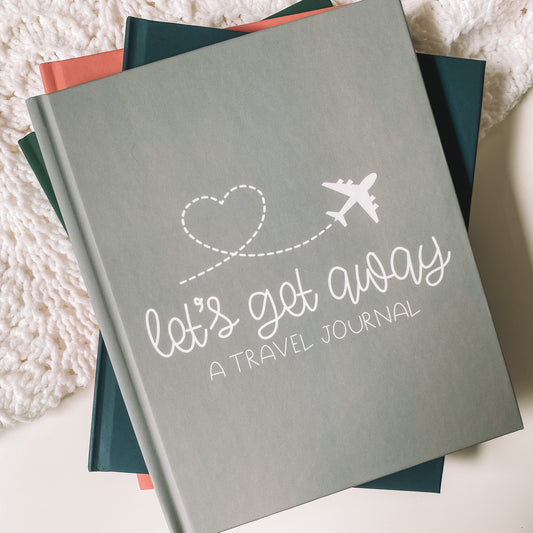 Keepsake travel journal with a soft, matte grey hardcover. White text reads Lets Get Away A Travel Journal with a graphic of a white airplane leaving a heart trail behind it.