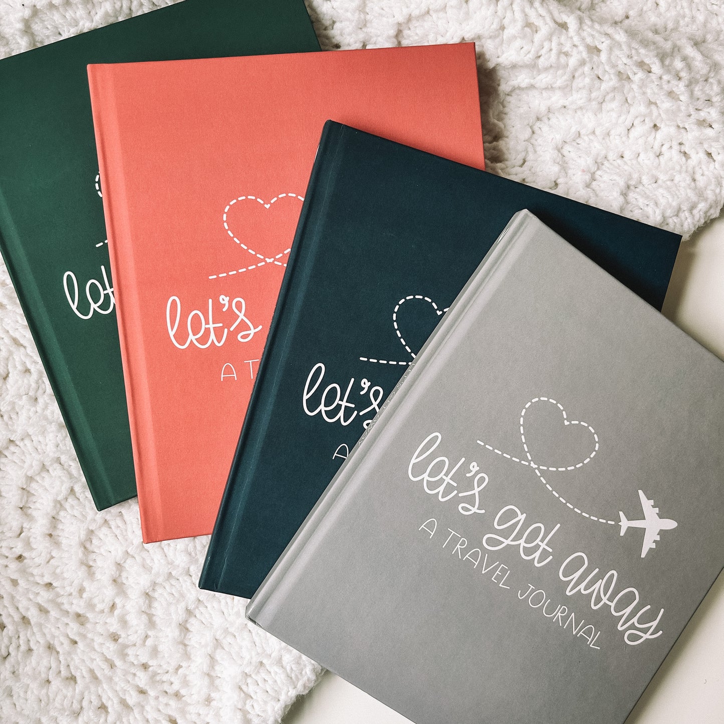 This keepsake travel journal is available in four colors which are forest green, navy blue, grey, and coral. All have white text that reads Lets Get Away A Travel Journal with a graphic of a white airplane leaving a heart trail behind it.