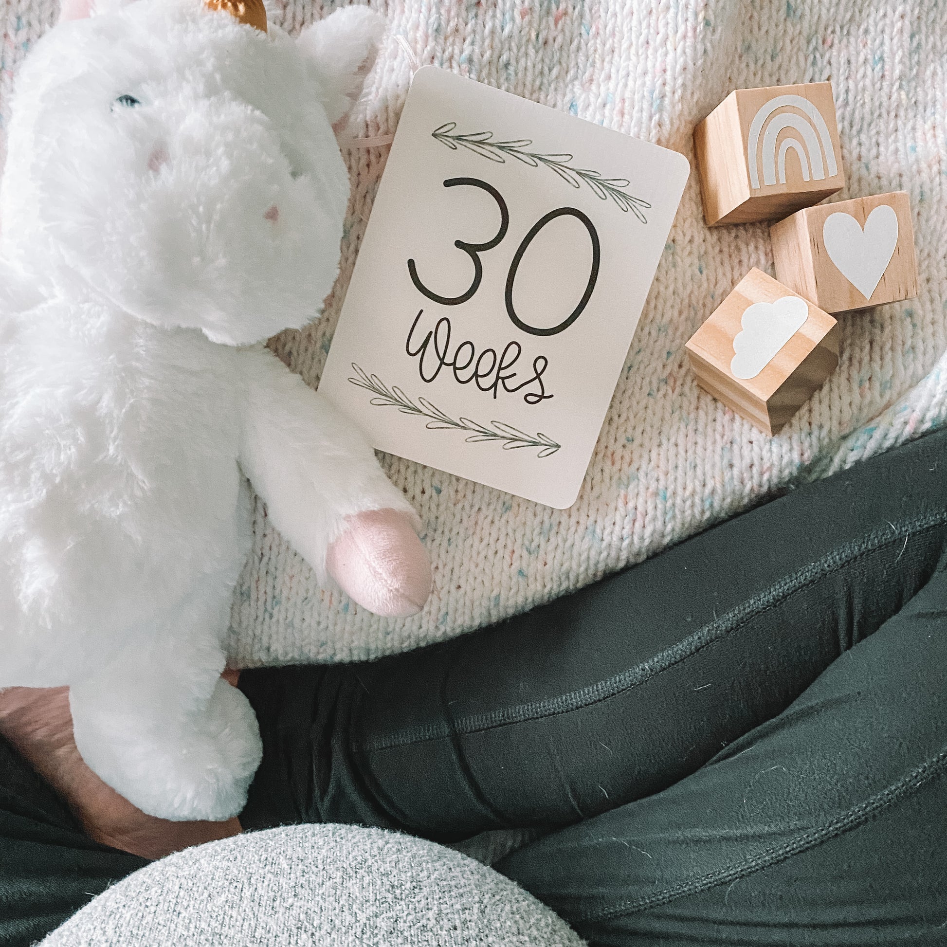 View of pregnant belly with a 30 week milestone card, unicorn stuffed animal, and blocks laid on a blanket in front.