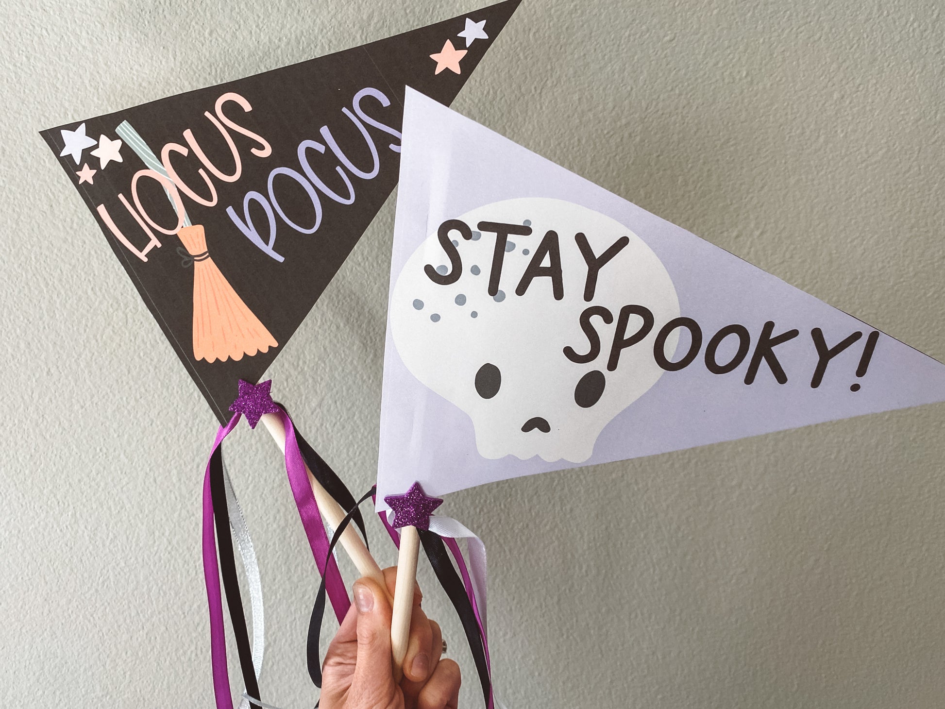 Hocus Pocus flag is black. Hocus is in light orange. Pocus is in light purple. Broom in the background and stars in two of the corners. Stay Spooky flag is light purple with black text and a cute white skull in the background.