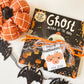 Halloween board book with ribbon wrapped around it and a happy halloween gift tag featuring three orange pumpkins and the gift giver's name at the bottom. Orange pumpkin and bat garland complete the flat lay.