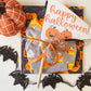 Happy Halloween pennant flag is white with orange text and three orange pumpkins below. Placed on top of a set of Halloween pjs and book. Orange pumpkin and black bat garland complete the flat lay.