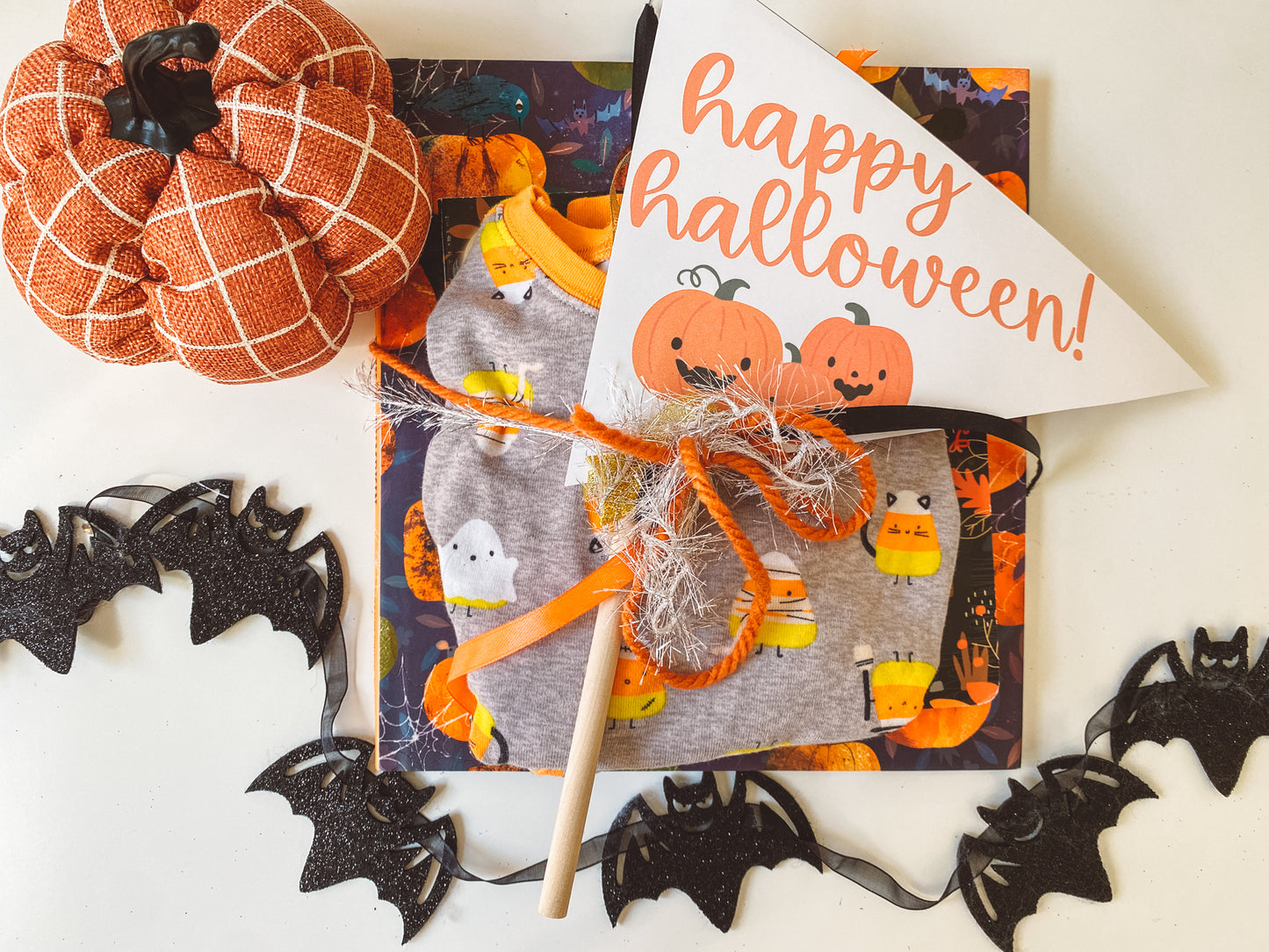 Happy Halloween pennant flag is white with orange text and three orange pumpkins below. Placed on top of a set of Halloween pjs and book. Orange pumpkin and black bat garland complete the flat lay.
