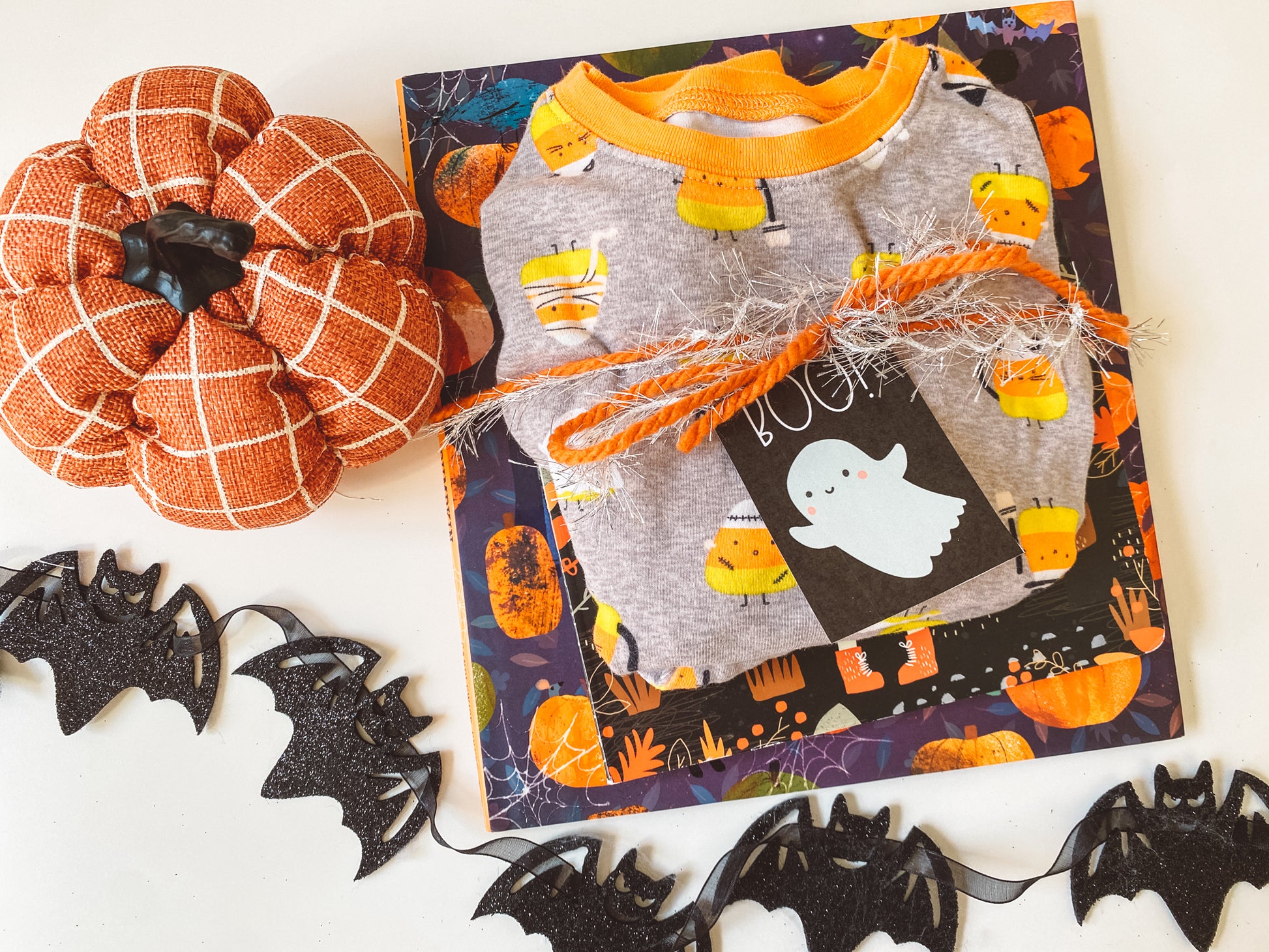 Book and Halloween pajamas wrapped up with yarn with a Boo gift tag attached. Orange pumpkin and black bats complete the flat lay.